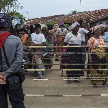 A crowd of people stand behind a fence while an armed officer watches them